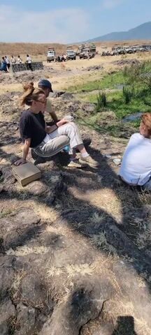 The video shows three people sitting on rocks near a lake on a sunny day. Two people are sitting together with a pizza box and cups nearby. The third person sits alone facing the lake with a thermos and other items beside her. The lake is bordered by grass and has ripples on the surface. A group of safari vehicles is parked in the distance. There are hills in the background.