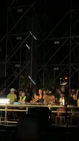 The video shows a nighttime outdoor event with a crowd gathered around a stage. A metal truss structure with lights is visible behind the stage. On the stage, there are several people operating DJ equipment. Some are wearing bright vests, suggesting they are part of the event staff. The crowd appears to be enjoying the music and atmosphere, with some individuals visible closer to the stage. The lighting is dynamic, with spotlights and illuminated vertical bars on the truss. The overall scene suggests a music festival or concert.