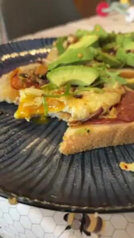 Here is a description of the video:

A close-up shot shows a dark blue plate with an irregular ridged surface. On the plate are two open-faced sandwiches. One has a bite taken out of it, revealing a runny egg yolk. Both sandwiches have toast as the base, topped with what appears to be slices of salami, a fried egg on one, and sliced avocado and a small amount of arugula on both. The background shows a white tablecloth with a honeycomb pattern. The lighting is natural.