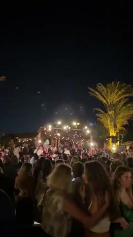 This is a vertical video shot at night outdoors. The scene appears to be a crowded outdoor venue, possibly a restaurant, bar, or party. There are many people, mostly young adults, gathered together. The lighting is dim, with string lights and spotlights illuminating parts of the scene. Several palm trees are visible, their fronds lit up by the lights. The camera is moving, creating a somewhat blurry and dynamic effect. The overall atmosphere suggests a lively and social event.