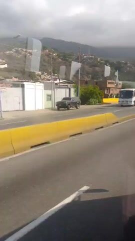 The video shows a view from inside a vehicle moving along a highway in what appears to be a mountainous region. The road is multi-lane with yellow painted lane dividers. In the first few seconds, a white bus is on the opposite side of the road. Later, two motorcyclists pass in the same direction. Buildings are visible in the background on the hillside. The sky is overcast. A white van-like vehicle then passes on the opposite side of the road.  The vehicle from which the video is being shot continues its forward movement.