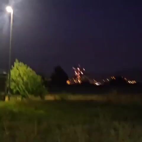 The video captures a nighttime scene, likely a fireworks display. In the foreground, a tall, lit streetlight stands to the left, casting a bright glow. A dense green bush is situated in the middle ground, partially obscuring the view beyond. The background reveals a dark, rolling landscape dotted with the warm lights of a town or city, suggesting an urban or semi-urban setting.

The main event is a vibrant fireworks burst in the distance, emitting bright red and white light against the dark night sky. The fireworks appear to be exploding over a hilly terrain, with numerous city lights visible below them. The scene is peaceful and celebratory, evoking a sense of occasion.

The time of day is clearly night, as indicated by the dark sky and the artificial lighting from the streetlight and the town. The weather appears to be clear, allowing for an unobstructed view of the fireworks. While no specific weather conditions are evident, the overall atmosphere suggests a pleasant evening for an outdoor event. The location cues, such as the general landscape and the style of lighting from the town, are consistent with a European setting, and specifically Mas Aliu, Spain. There is no visible text in the media file.