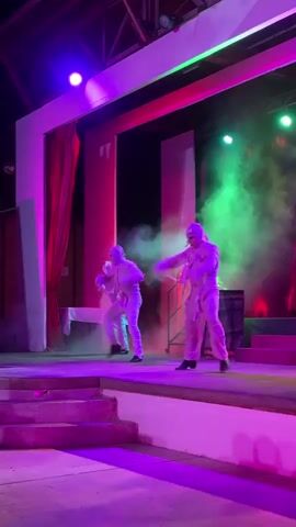 The video shows a performance on a stage. There are several people dressed in white, some appearing to be mummies or ghosts, dancing and moving in a dramatic fashion. One individual in a red cloak stands on a raised platform at the back of the stage, possibly playing a character like a queen or a sorceress. There is smoke or fog on stage, illuminated by colored lights in pink, purple, and green. A barrel with what appears to be fire or light emanating from it is visible on stage. The overall scene suggests a theatrical or entertainment performance, possibly with a spooky or dramatic theme.
