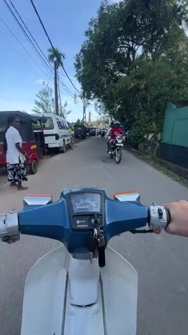 A first-person perspective shows a person riding a white and blue Honda scooter down a narrow, paved road in what appears to be a tropical location.  The road is lined with parked vehicles and people, many of whom are wearing white clothing.  There are also red and yellow Buddhist flags and strings of white lanterns hanging overhead, suggesting a religious or cultural event.  The sky is blue with some clouds, and palm trees and lush vegetation are visible along the roadside.  The rider is moving slowly, weaving through the crowd.