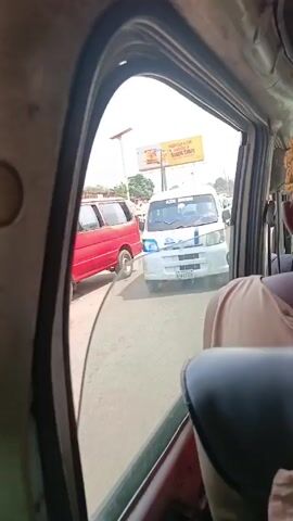 The view from inside a moving vehicle shows a woman in the passenger seat looking out the open window. Several small vans, most of them white, are lined up on the road outside. A billboard advertising "Double Cubes" is visible in the background. A man walks across the road between the vans. The vehicle appears to be moving slowly in traffic.