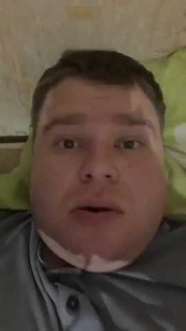 A close-up video shows a man's face. He is speaking to the camera, and his mouth is open as if he is talking. The man appears to be lying down, possibly in bed, as evidenced by the soft, green patterned surface visible behind his head. His skin is pale, and he has dark hair. There are areas of lighter skin on his forehead and cheeks, possibly due to lighting or a skin condition. The background is blurred, suggesting the focus is entirely on the man's face. The man is wearing a gray shirt.