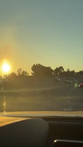 The video shows a view from inside a vehicle traveling on a highway at sunset. The sun is low in the sky, casting a warm, golden light and creating lens flare. Other vehicles are visible on the highway, some in motion and some appearing to be stopped in traffic. Trees and hills are silhouetted against the sky in the distance. The interior of the vehicle, including a dashboard element, is partially visible in the foreground. The overall scene depicts a typical commute or travel experience during dusk.