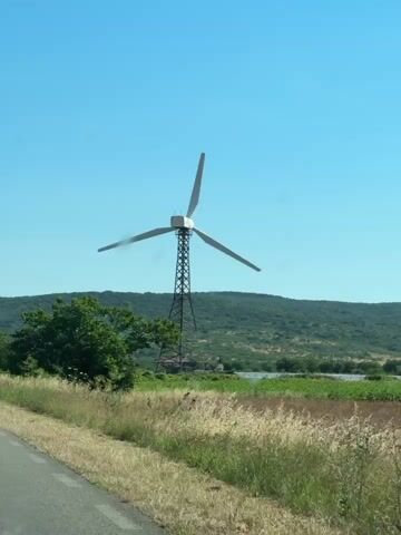 This video captures a view from a moving vehicle on a sunny day. The main subject is a tall wind turbine with three blades. In the foreground, a grassy roadside with some dry stalks and a portion of an asphalt road is visible. To the left, a green tree stands out against a backdrop of rolling, tree-covered hills under a clear blue sky. As the vehicle moves, a triangular road sign indicating a sharp bend or turn appears to the left of the turbine. The turbine's blades are angled upwards and outwards, and the structure appears to be a lattice tower. The landscape suggests a rural or semi-rural environment.
