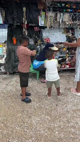 In Tambon Krasom, Thailand, two young boys and at least two other individuals are captured in a still image. The setting appears to be an outdoor market stall, densely packed with an assortment of merchandise, including necklaces, bracelets, and other trinkets displayed on racks. A dark tarp forms the backdrop of the stall, with various items hanging from it.

The primary activity seems to be a moment of interaction, possibly play, between the two boys. One boy, wearing a tan t-shirt and dark shorts, is holding a yellow object, perhaps a toy, towards a seated figure. This seated figure, a woman in a blue shirt and headscarf, is leaning forward, partially obscured. The other boy, in a white and yellow t-shirt and light shorts, stands facing away from the camera, also holding a yellow object. Another person, wearing a striped dress, stands to the right of the frame, their hand extended. A younger boy in a maroon shirt and blue cap is also visible to the right.

The ground is covered in small pebbles. The lighting suggests it could be daytime, possibly overcast, given the lack of harsh shadows. The overall mood seems casual and everyday, with the children engaged in their activities amidst the bustling market environment. No visible text is present in the frame.