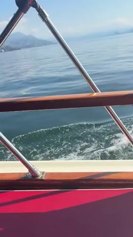 The video captures a scenic boat ride on a bright, sunny day, presumably on a lake or large body of water in Binda, Italy. The perspective is from the stern of a moving boat, showcasing the tranquil surroundings.

The initial shots frame the view over a wooden and metallic boat railing, looking back at the foamy white wake generated by the vessel. Distant mountains or hills rise from the horizon under a clear, bright blue sky, their forms slightly softened by a gentle haze. The water appears calm, with ripples and glints of sunlight reflecting off its surface.

As the video progresses, the camera pans to reveal more of the boat's wake and the expansive water. Around the 0:05 mark, a portion of what appears to be an Italian flag, with its green, white, and red vertical stripes, becomes visible near the boat's edge, fluttering gently. The shoreline comes into view, displaying lush green vegetation and what seem to be distant buildings along the base of the landmasses. The bright sun casts a warm glow over the entire scene, creating sparkling reflections on the water's surface.

The video ends with a wide shot encompassing the boat's trailing wake, the vast expanse of water, and the distant, tree-covered hills under a brilliant, cloudless sky. The activity depicted is simply the boat traveling through the water, offering picturesque views. No people are explicitly visible, and no text appears in the video. The time of day is clearly daytime, characterized by strong sunlight and clear weather.