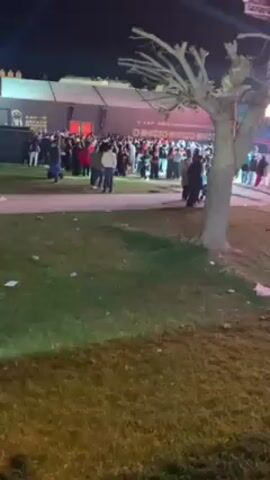 At night, a large crowd of people gathers outside a building, likely an event venue, as evidenced by banners and signage. The scene is viewed from a low angle across a grassy area littered with scattered debris. The crowd consists of individuals of various ages and appearances, some standing in groups, others walking around. Lighting suggests a celebratory or public event atmosphere. A barren tree stands prominently in the foreground, slightly obscuring the view of the crowd. The building behind the crowd has logos and words that are partially illegible but seem event related.