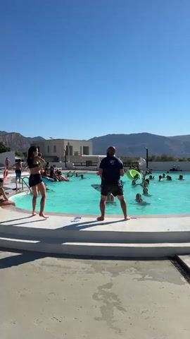 A man and a woman are dancing on the edge of a swimming pool. The woman is wearing a black bikini top and black shorts, and the man is wearing a blue t-shirt and black shorts. Several people are swimming and playing in the pool behind them. In the background, there are mountains and a building. The sky is clear and blue.