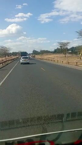 The video shows a view from inside a vehicle driving on a two-lane asphalt road. A white car is driving in front of the vehicle in the same lane, and a truck is further ahead. On the opposite side of the road, there is oncoming traffic. The road has a guardrail on the right side, and the landscape on both sides is primarily dry and grassy with scattered trees and utility poles. The sky is blue with scattered white clouds. The overall scene is a typical daytime road setting in a rural or semi-rural area.
