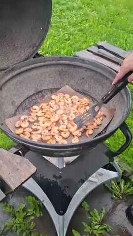 A close-up shot shows shrimp being cooked on a grill. The shrimp are arranged on a piece of parchment paper placed on a grill grate. A person's hand, wearing a dark jacket, is using a black spatula to turn the shrimp. The grill is open, and steam is rising from the shrimp. The background is blurred green grass and a wooden surface. The overall scene suggests outdoor cooking.