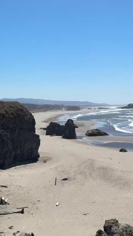 The video shows a wide view of a sandy beach under a clear blue sky. Large rock formations rise from the sand and jut out into the ocean. Waves crash against the shore and the rocks, creating white foam. In the distance, the coastline curves, with more rock formations and mountains visible on the horizon. A few small figures, likely people, can be seen walking on the beach near the water's edge. The sand appears to be wet closer to the water and drier further up the beach. Some driftwood and debris are scattered on the sand. The overall impression is a natural, undeveloped coastal landscape.