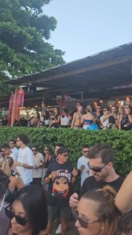 The video depicts a crowd of people at an outdoor event, likely a party or concert. Many attendees are filming or taking photos with their phones. The setting appears to be a beachside or near-beach location, with some greenery in the background. A structure, possibly a stage or bar area, is visible with people both behind and in front of it.  Most of the people are wearing casual clothing and sunglasses. The crowd is dense, indicating a popular or well-attended event. The video seems to be shot from within the crowd, providing a first-person perspective of the event.