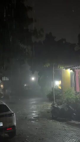 This media file captures a scene at night during a heavy rainstorm. The setting appears to be a residential area in Baan Bang Rak, Thailand, suggested by the tropical foliage and architectural style of the buildings.

The primary subjects are the elements of the storm and the surrounding environment. Rain is falling heavily, creating a hazy, misty atmosphere. The ground is wet and reflects the dim lighting. In the foreground, the rear of a white SUV is visible. To the right, a yellow building with a pink trim is illuminated by interior lights, with plants and a covered motorcycle parked outside. Trees and dense foliage dominate the background, silhouetted against the dark night sky.

The main activity is the downpour. A person wearing a bright red raincoat is walking away from the camera down the wet path. The time of day is clearly night, indicated by the darkness and the artificial lights. The weather is a significant feature, with the rain creating a dramatic and atmospheric mood. The overall impression is one of a tropical night disrupted by a powerful rain shower. There is no visible text in the media file.