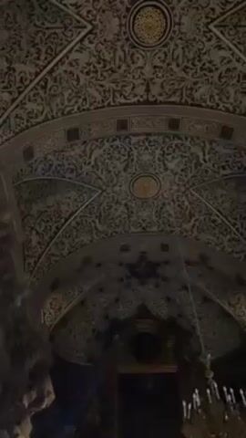 The camera pans down in a dimly lit church, starting with a focus on the intricately decorated ceiling, then moving down to reveal a golden altar with a crucifix, surrounded by candelabras. The altar is adorned with sculptures and artwork. The overall atmosphere is ornate and sacred.