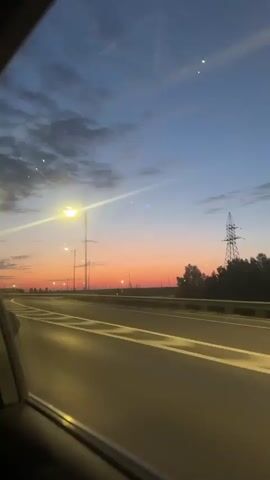 A video shot from inside a moving vehicle shows an empty highway at dusk. The sky is a gradient of orange near the horizon, fading into blue with some clouds above. Streetlights illuminate the highway. An electricity tower is visible in the distance. Reflections appear on the car window.