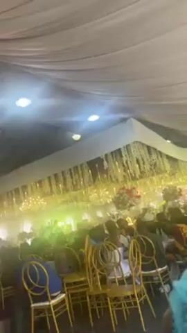 The video shows a view of an indoor gathering. A crowd of people sit on gold-colored chairs beneath a draped white ceiling adorned with lighting and floral decorations. Someone in a pink shirt walks through the scene. The lighting is bright, creating a vibrant atmosphere.