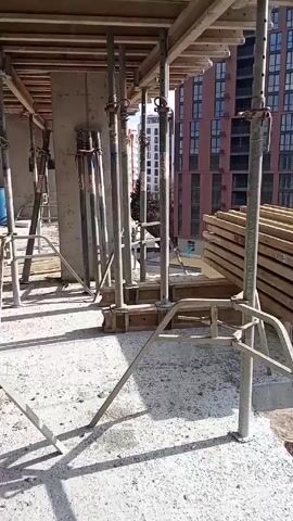The video shows a construction site with a stack of wooden beams being lifted by a crane. The beams are stacked horizontally and secured with cables. The crane is lifting the beams from a higher level of the construction site. Below, there is a partially constructed building, construction equipment, and a porta potty. Several other buildings are in the background. The ground level is largely dirt and debris. The sky is bright and sunny.
