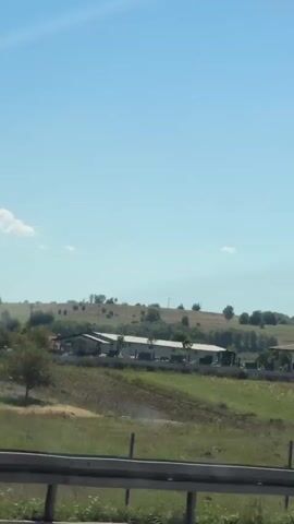 This video appears to be a dashcam recording from a vehicle. The camera is angled forward and slightly upwards, capturing a rural landscape under a clear blue sky with a few wispy clouds. In the foreground, a metal guardrail is visible, partially obscuring the view. Beyond the guardrail, a grassy field stretches out, dotted with rocks and small bushes. A wire fence runs across the field. In the middle ground, a low-rise building complex with a tiled roof is visible, along with some trees and agricultural land. Further in the distance, rolling hills are covered in sparse vegetation. A prominent white building with a pointed steeple, likely a church, stands out against the landscape. Another vehicle, a dark-colored SUV, is seen driving in the opposite direction in the right portion of the frame. The camera pans slightly to the right as the video progresses.