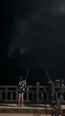 In the video, a young woman and a young man stand on a bridge or embankment at night, lighting fireworks. The woman, positioned on the left, wears a dark-colored shirt with white floral patterns and short shorts. The man, on the right, is dressed in a dark jacket and shorts. Both hold sticks, from which the fireworks are launched into the dark sky. The fireworks explode into a variety of colors including pink, white, and green, creating a visual spectacle against the night sky. Below them is a blue scooter parked. The backdrop features dark water and distant lights, suggesting a waterfront setting.