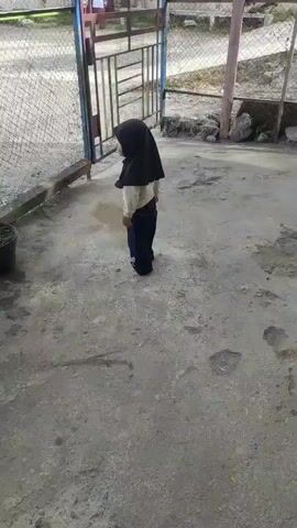 A young child wearing a black hijab and a light-colored shirt with dark pants stands in front of a metal gate. The child is facing away from the camera and appears to be interacting with the gate, possibly trying to open it. The ground is concrete, with some patchy areas and a small puddle. The gate is partially open, revealing a glimpse of what appears to be a schoolyard or outdoor area with other children and buildings in the background. The scene is outdoors, and the lighting suggests it is daytime.