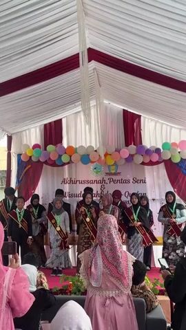 A group of students, likely on a stage, are participating in what appears to be a graduation or award ceremony. They are dressed formally, with some wearing sashes and medals. The stage is decorated with balloons and a banner that reads "Akhirussanah, Pentas Seni & Wisuda Tahfidz Al-Qur'an". Several adults are seated in the foreground, some holding up phones to record the event. The setting seems to be outdoors, under a large tent.