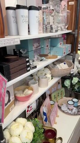 The video shows shelves stocked with various merchandise in a retail setting. On the top shelf, there are white travel mugs, clear glass containers, and other decorative items. The second shelf displays more mugs, boxes of kitchenware, and a woven basket. The third shelf contains a pink bowl filled with white, oval-shaped objects, several textured mugs, and decorative trays with cartoon characters. The bottom shelf features a large planter with artificial white flowers, a metal tray with two small red bowls, and additional decorative items. The overall environment appears to be a gift shop or home goods store. There are no people visible in the video.