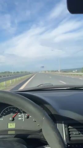 The video offers a first-person view from inside a car traveling on a multi-lane highway under a partly cloudy sky during daytime. The dashboard is visible in the foreground, showing a portion of the steering wheel on the left. The speedometer, marked from 0 to 200 km/h, has its needle resting at 0 km/h, while the odometer displays "13090.4" kilometers. Another digital readout shows "1827".

The camera initially focuses on the road ahead, showing the asphalt, guardrails, and numerous streetlights lining the highway. As the video progresses, the view pans slightly upwards to reveal a blue highway sign. The sign, located overhead, provides directional information. It reads "M7", "ОЧЕР" (Ocher) with a straight arrow, and "УСТЬ-СЫНЫ" (Ust-Syny) with a right-pointing arrow, followed by "300м" (300 meters). In the distance, scattered trees and what appear to be rural buildings or houses are visible on the horizon. The overall scene depicts a clear, open road.