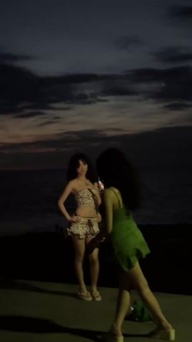 Two women are on a beach at dusk. The sky is dark with streaks of orange on the horizon. One woman is wearing a strapless leopard-print top and matching shorts, as well as sandals. The other is wearing a short green dress with thin straps. The woman in the green dress is facing away from the camera and holding a phone up to take a photo of the woman in the leopard print. The woman in leopard print is posing with her hand on her head. The sea is visible in the background.