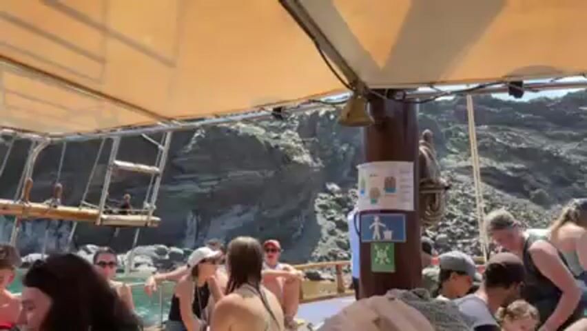 The video shows a group of people on a boat, seemingly enjoying a sunny day. They are sitting and standing, some looking out at the rocky coastline in the background. The boat has a canopy for shade and a wooden mast with a bell hanging from it. There are signs and ropes attached to the mast.