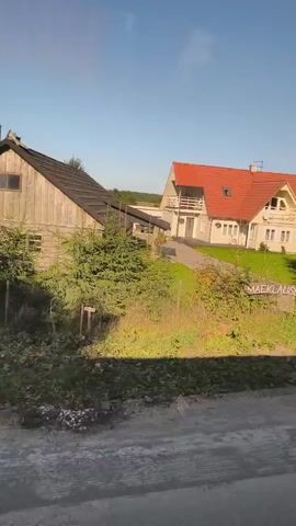 The video captures a view from a moving vehicle passing by a rural setting. The scene includes a house with a red roof, a smaller wooden structure, a driveway, a gate, and lush greenery. A sign with the text "MAEKLAUSE" is visible in front of one of the buildings. The sun is shining, casting shadows, and the sky is clear and blue. The overall impression is of a sunny day in a residential area surrounded by nature.