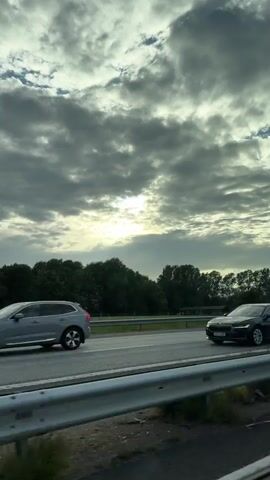 The video shows a view from a car traveling on a highway. The sky is cloudy with the sun partially visible through the clouds, casting rays of light. On the side of the highway, there are trees and a few buildings, possibly houses or barns, in the distance. A metal guardrail is visible in the foreground. The road is paved and appears to be a multi-lane highway. The overall impression is of a cloudy day with the sun trying to break through, seen from a moving vehicle.