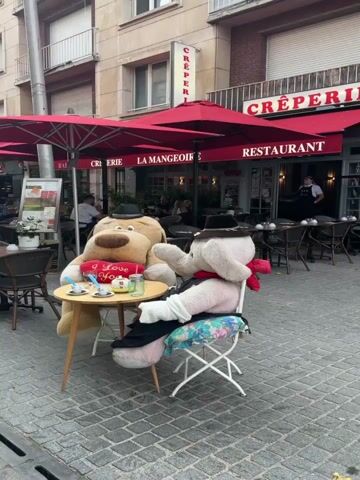 The video shows two stuffed animal characters, a large brown dog and a white elephant, sitting at a small round table outside a restaurant. The dog, dressed in a black hat and holding a red heart that says "I Love You," faces the elephant. The elephant, also wearing a hat and a red scarf, is seated on a floral cushion and appears to be talking to the dog. On the table between them are two small cups, a teapot, and a glass of lemonade. In the background, the restaurant, identified as a creperie and restaurant, has red awnings and tables with chairs set up outside. Several people are visible inside the restaurant and at other tables on the patio. The ground is paved with cobblestones, and a sidewalk stretches along the side of the building, lined with more outdoor seating. The overall setting appears to be a European street scene on a moderately overcast day.