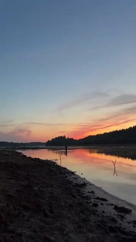 A person stands in shallow water by a riverbank at sunset. The sky is a gradient of blue and orange, with clouds reflecting the colors of the setting sun. The water reflects the sky and the distant treeline. The foreground shows a sandy bank littered with debris. The video pans slightly to the right, following the movement of the person.