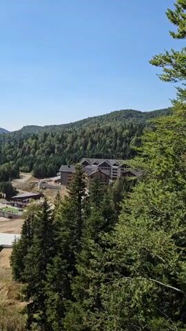 A drone flies over a forest towards a multi-story building with a dark roof. The building appears to be a hotel or resort, with multiple balconies and windows visible. The surrounding landscape is characterized by rolling green hills covered in dense trees. The sky is clear and blue, indicating fair weather.