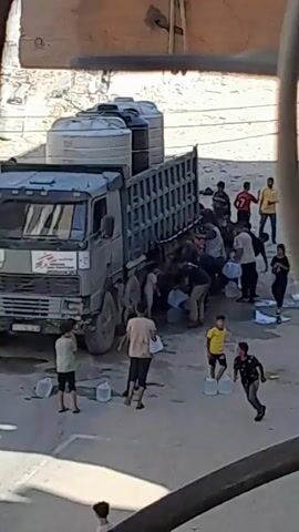 A group of people, including children, are gathered around a large truck with a high-sided bed. The truck has several large water tanks on its bed. Some individuals are reaching into the back of the truck, and others are holding large plastic containers, appearing to fill them with water from the truck. A logo resembling "MSF" is visible on the side of the truck. The ground appears to be dirt and rubble. Some people are running or walking, carrying the filled containers. The scene appears to be outdoors during the daytime.