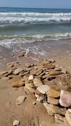 The video shows a beach with waves crashing on the shore. There are many rocks and stones scattered on the sand. The water is blue-green, and the sky is clear.