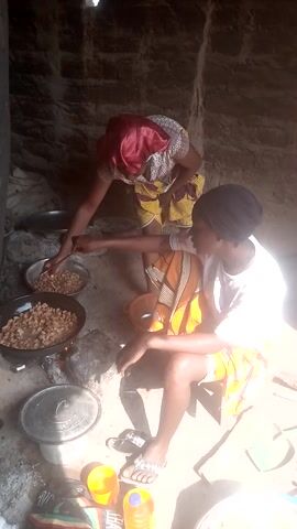 Two women are engaged in food preparation in what appears to be an outdoor or semi-outdoor cooking area. The setting features a dirt ground and a rough stone or mud wall in the background, illuminated by bright daylight casting strong shadows.

One woman, positioned centrally, wears a white t-shirt, a dark headwrap, and a vibrant orange and yellow patterned skirt. She is leaning over two dark metal pots, actively stirring the contents of one with a stick or spoon. The pots are placed on a traditional cooking setup, likely over a fire that is not fully visible. The contents of the pots and a nearby large metal tray appear to be roasted peanuts or groundnuts.

To her left, another woman is partially visible, wearing a patterned dress and a red head covering. She is reaching with her hand into a smaller metal bowl, also containing the same type of nuts, suggesting she is sorting or preparing them.

Around the cooking area, several items are scattered on the ground: a yellow plastic bottle, a yellow plastic cup, and a colorful piece of fabric or bag with the partial text "HAR" visible. In the distant background, a green shovel can be seen leaning against a surface. The scene captures a moment of collaborative food processing, indicative of daily life in a rural or traditional setting.