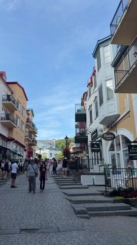 A group of people are walking down a cobblestone street lined with colorful buildings. Some of the buildings have balconies with balconies. There are shops on the ground floor of the buildings. The sky is blue with some clouds.