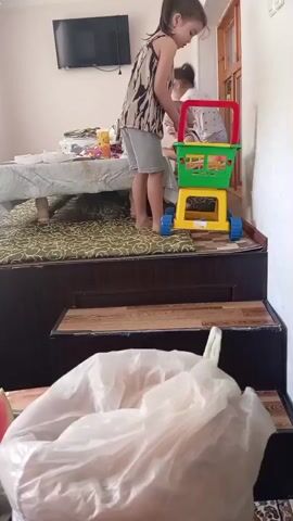 Two young children are playing with a toy shopping cart indoors. A girl with dark hair tied in a ponytail, wearing a patterned top and gray shorts, stands and interacts with the cart. She is barefoot. Another child, with lighter hair and wearing a white shirt, is partially visible in the shopping cart, possibly being pushed or playing with items inside. The toy cart is colorful, with a yellow base, green basket, and red handle. It has blue wheels. The children are on a raised platform covered with a green patterned rug. There is a table covered with a white cloth to the left, with various items on it, including what appear to be children's toys and containers. A television is mounted on the wall in the background. In the foreground, a large, translucent plastic bag is visible, obscuring the lower part of the scene. The environment appears to be a living room or play area.