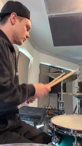 The video shows a young man in a black hoodie and a black baseball cap playing a drum set in what appears to be a recording studio or practice room. He is focused and intense, rhythmically striking the snare drum and cymbals with his drumsticks. The room is well-lit, with acoustic panels visible on the ceiling. Other drums and an electronic percussion pad are visible nearby. The camera remains stationary throughout the short clip, capturing the drummer from the chest up.