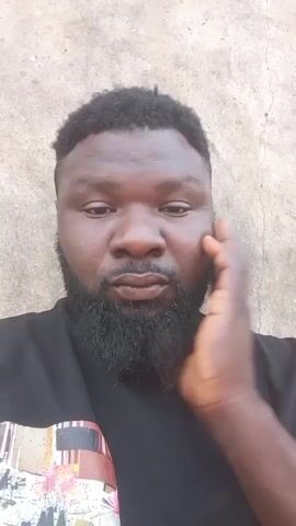 A man is looking at the camera while touching his beard. He has dark skin, a short afro-style haircut, and a full black beard. He is wearing a black t-shirt with a design on it. He is standing in front of a light grey textured wall.
