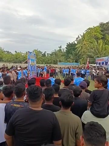 A medium shot captures a crowded outdoor event. In the foreground, a group of men, primarily viewed from behind, are looking towards a raised stage. On the stage, several individuals in blue shirts are gathered around a podium, some interacting with each other. Behind the stage, a banner with text and images is displayed. A white tent is visible in the background to the right, with more people milling around it. The environment appears to be a grassy field, with trees and a gentle slope in the background. The sky is overcast. A young child is being carried on someone's shoulders in the mid-ground, also facing the stage. The overall impression is of a community gathering or a public event.
