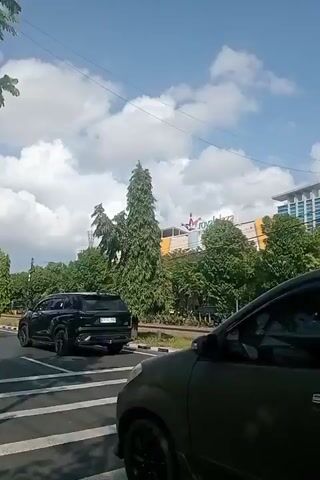 A daytime, outdoor shot captured from a moving vehicle, likely a car. The camera perspective is looking out the side window. In the foreground, the back of a dark-colored SUV is visible, with a license plate that reads "B 8063 BGX". The SUV is on a paved road, with a white line marking the lane. Beyond the road, there are train tracks, bordered by grass and a low curb. In the middle ground, a tall, slender green tree stands prominently. Behind the tree, a large, modern building complex is visible. The building has a multi-colored facade, with sections of yellow, orange, and blue. Signs are visible on the building, but the text is not legible. Several other vehicles are parked or driving in the background. Trees and utility poles are scattered throughout the scene. The sky is blue with scattered white clouds. The overall impression is of a suburban or semi-urban environment on a clear day.