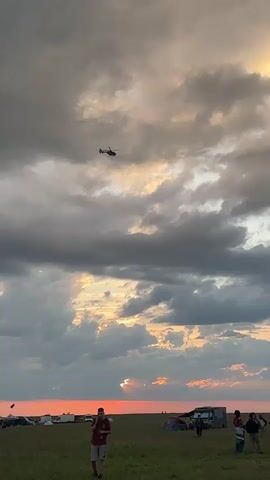 The video shows a sunset over a field with a crowd of people. A helicopter flies overhead. There are several RVs and tents set up in the field. The sky is cloudy and the sun is setting on the horizon.