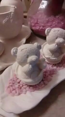 THE VIDEO SHOWS A CLOSE-UP, SLIGHTLY BLURRY SHOT OF A DISPLAY OF WHITE CERAMIC TEDDY BEAR FIGURINES. THE FIGURINES ARE ARRANGED ON A WHITE, LEAF-SHAPED PLATTER WHICH IS FILLED WITH SMALL, CRUSHED PINK STONES. THERE ARE AT LEAST THREE TEDDY BEAR FIGURINES VISIBLE IN THE FOREGROUND AND MIDDLE GROUND, WITH A FOURTH PARTIALLY VISIBLE FURTHER BACK. THE TEDDY BEARS ARE DETAILED, WITH TEXTURED FUR AND SMALL, EXPRESSED FACES. ONE TEDDY BEAR IN THE FOREGROUND IS HOLDING A SMALL, HEART-SHAPED OBJECT. IN THE BACKGROUND, A LARGE GLASS VASE OR CONTAINER FILLED WITH MORE PINK STONES IS VISIBLE, ALONG WITH A RED CANDLE OR OBJECT. TO THE FAR LEFT, A PORTION OF ANOTHER WHITE CERAMIC OBJECT, POSSIBLY A TEAPOT OR VASE, IS SEEN. THE LIGHTING IS SOFT AND APPEARS TO BE INDOOR. THE OVERALL IMPRESSION IS A DECORATIVE DISPLAY, POSSIBLY FOR A CELEBRATION OR AS A TABLE CENTERPIECE.
