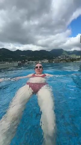 A woman is swimming in a rooftop infinity pool. She is wearing swimming goggles and a red swimsuit. She is facing the camera, floating on her back and paddling her arms and legs. In the background is a mountain range, a city with buildings and a bridge. The sky is partly cloudy. The water in the pool is clear and blue with small ripples.